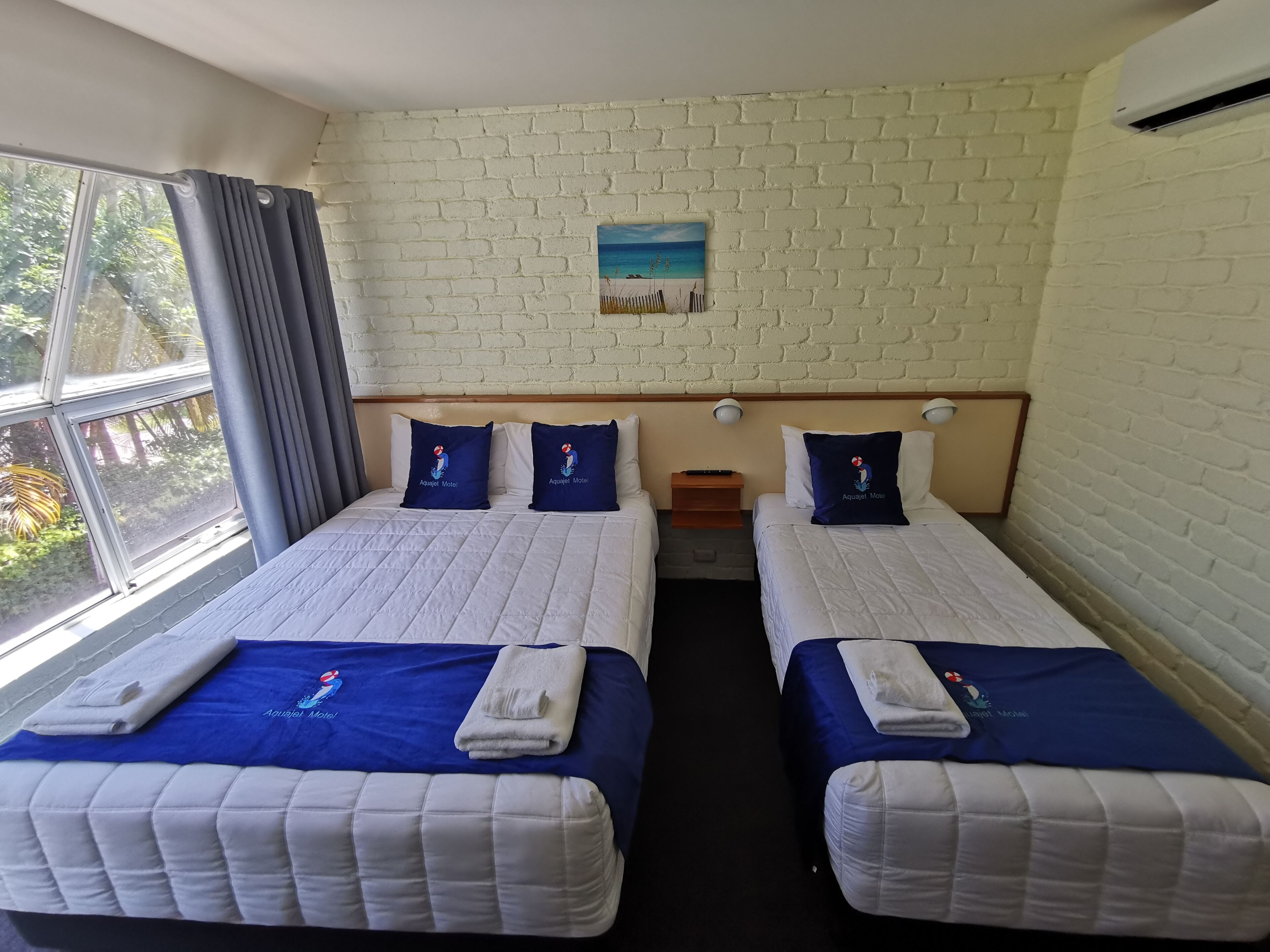 A hotel room with two beds, a brick wall, a painting of a beach, and a window with curtains.