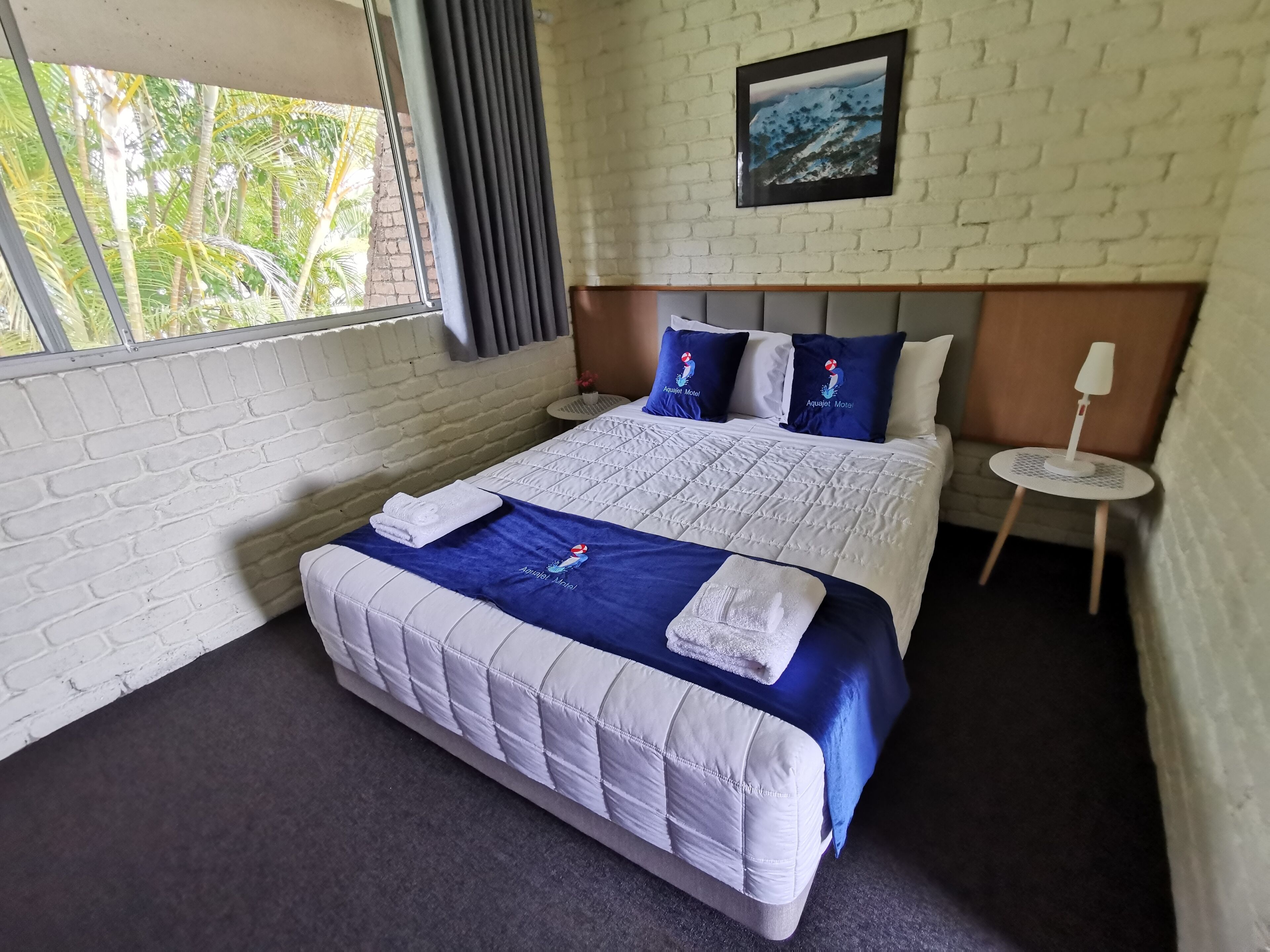 Una habitación de hotel con cama, ventana, un cuadro en la pared y una mesita con lámpara.