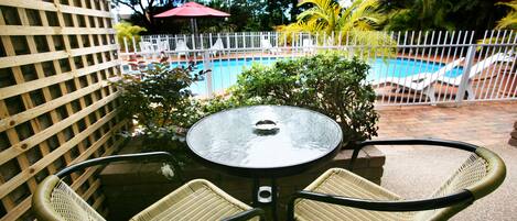 A patio area with a round table and two wicker chairs, a pool, and a white fence.