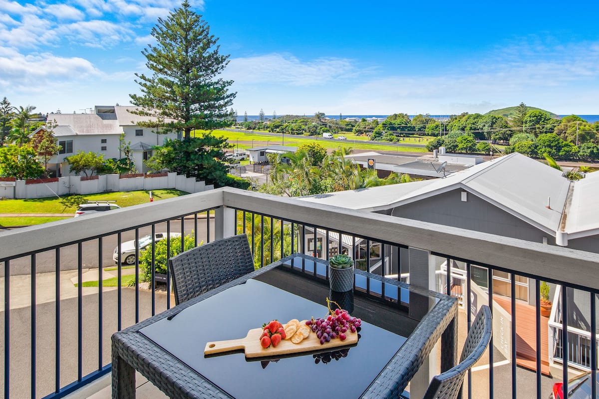 A balcony with a glass table, a wooden cutting board with fruit, and a view of a residential area with houses and greenery.