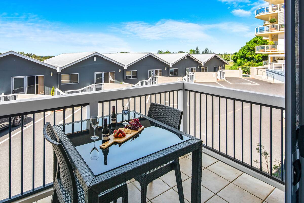 A balcony with a table set for a meal, a car parked in the background, and a row of similar buildings in the distance.
