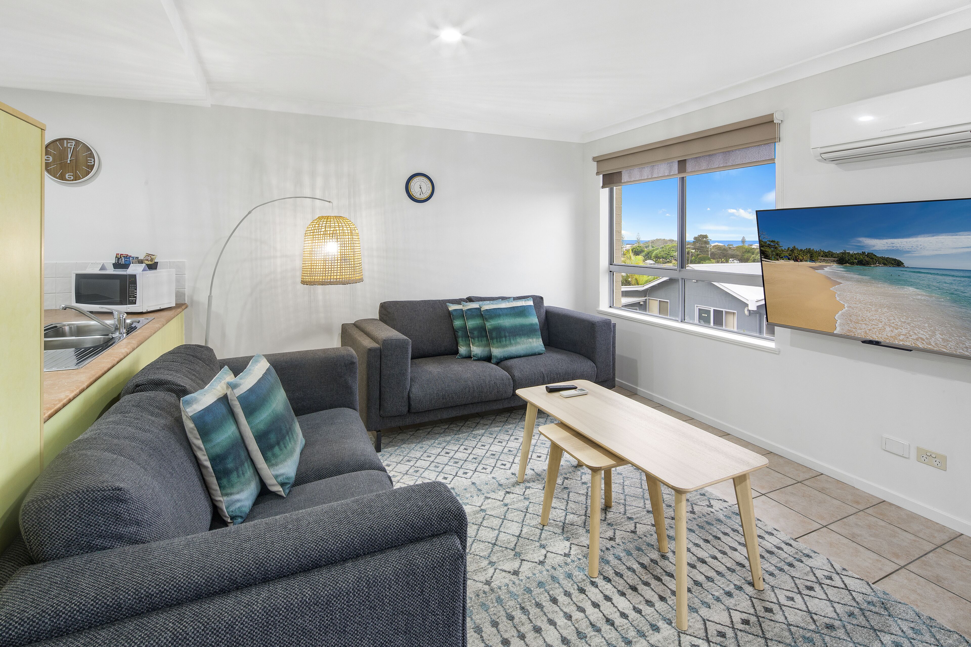 Een moderne woonkamer met een grijze bank, een houten salontafel en een groot raam met uitzicht op het strand.