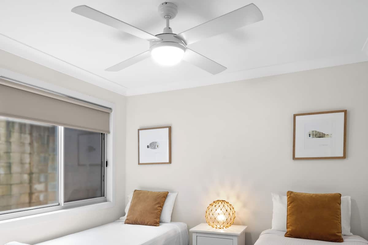 A hotel room with two single beds, a ceiling fan, a window with blinds, and two framed pictures on the wall.