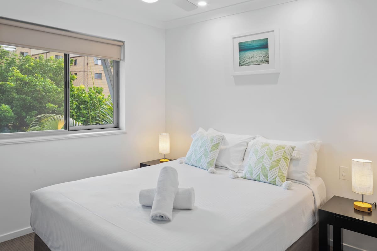 A neatly made bed with a rolled towel, a ceiling fan, a bedside lamp, and a window with a view of greenery outside.