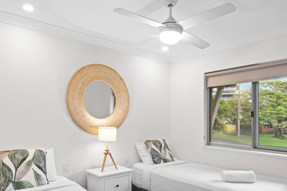 A hotel room with two beds, a ceiling fan, a round mirror, a bedside lamp, and a window with a view of greenery.