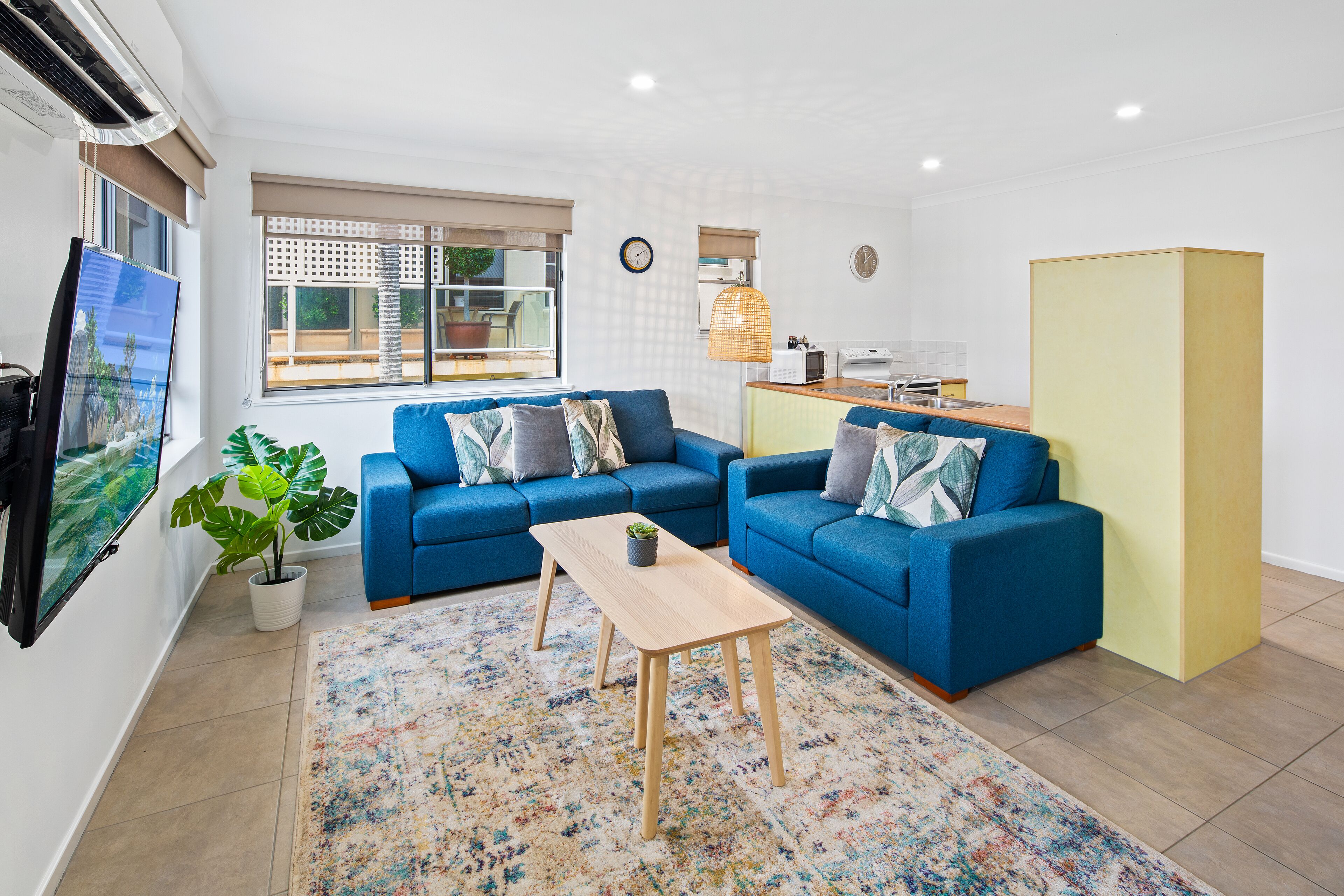 A modern living room with two blue sofas, a wooden coffee table, a potted plant, and a large TV screen.