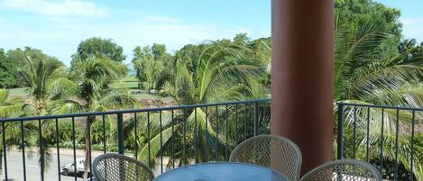 A balcony with a round table and wicker chairs, overlooking a green landscape with trees and a road.