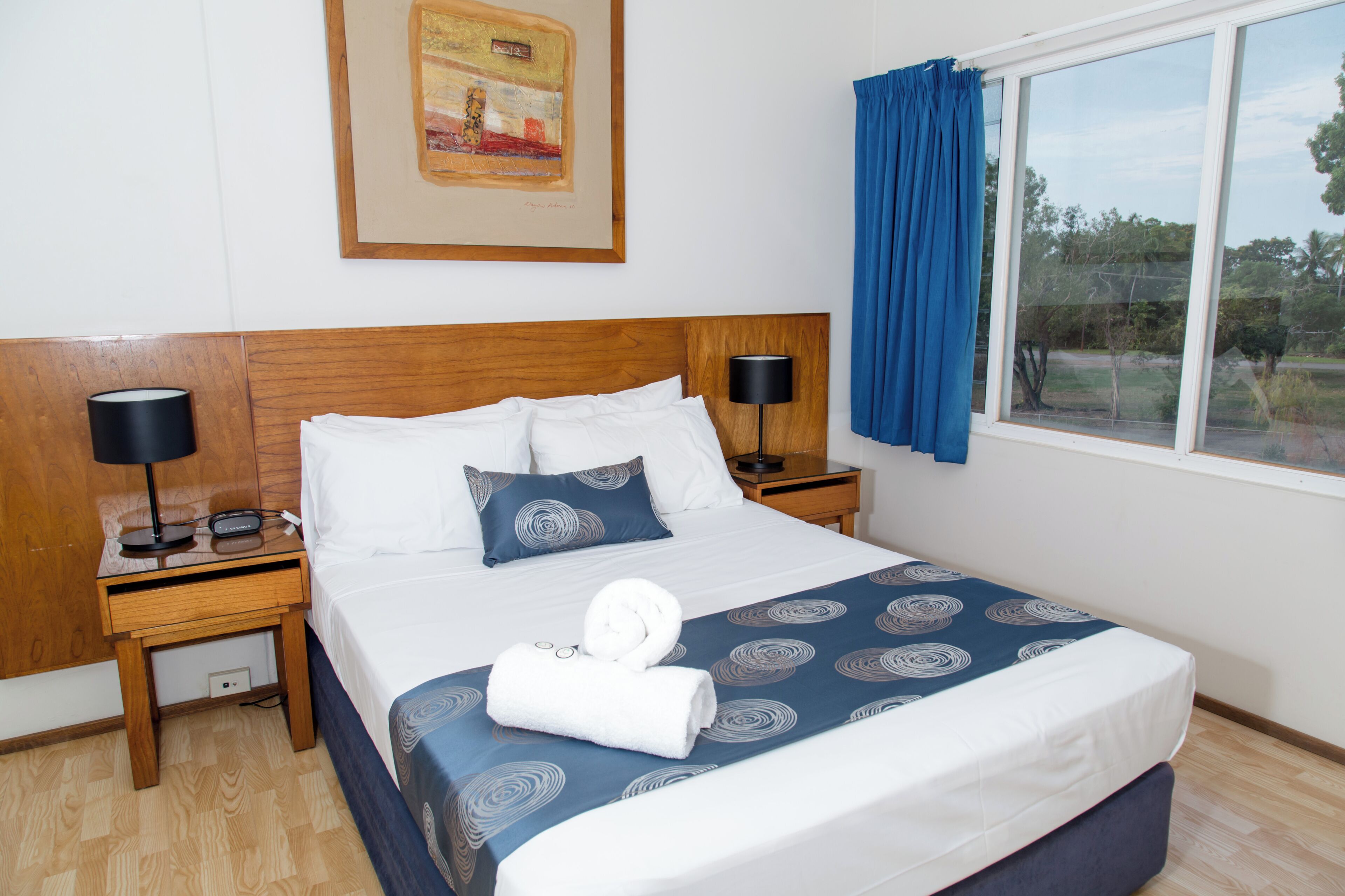 A neatly made bed with a blue and white patterned bedspread, two bedside lamps, a framed picture on the wall, and a window with blue curtains.