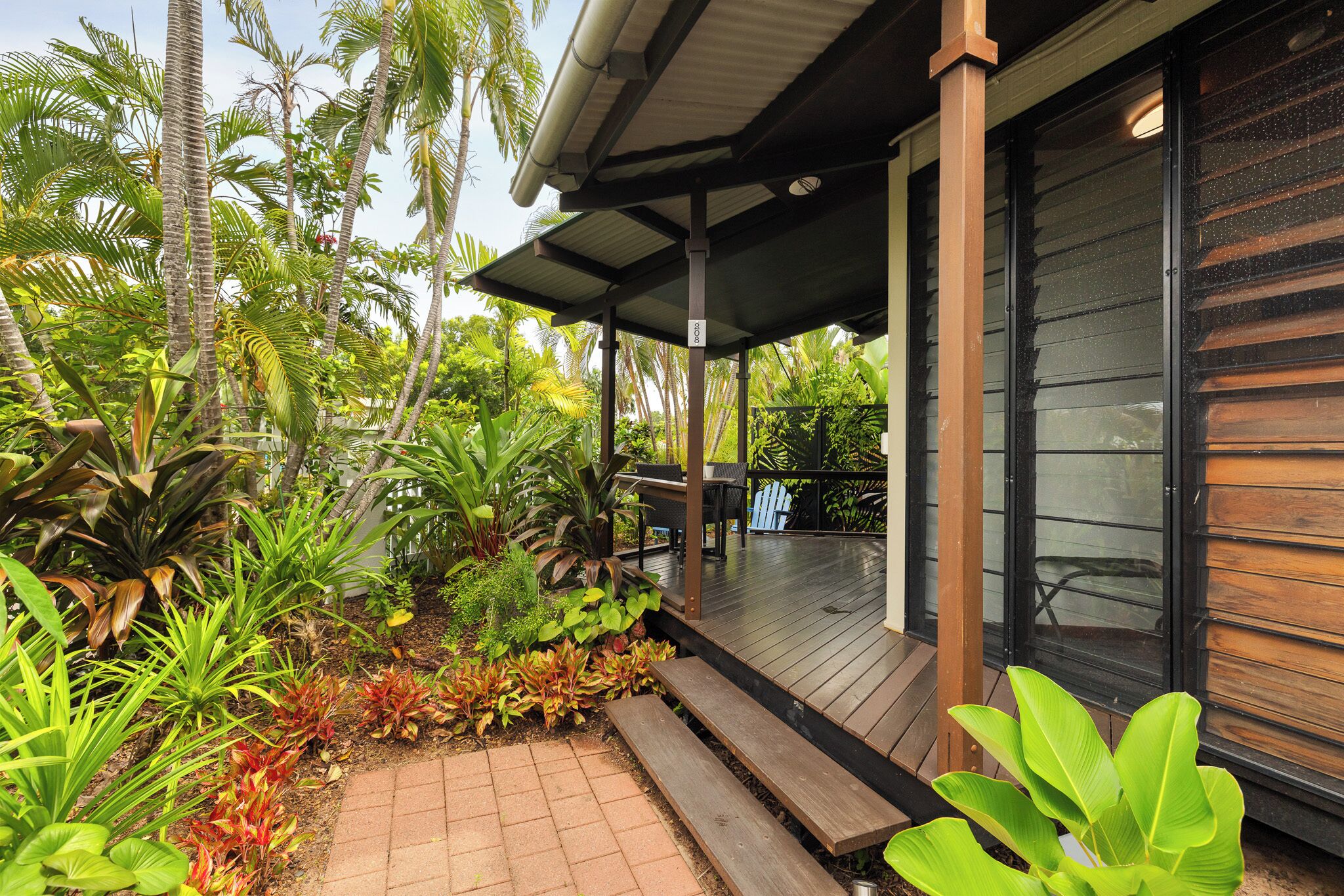 A covered outdoor area with a wooden deck, surrounded by tropical plants and palm trees.