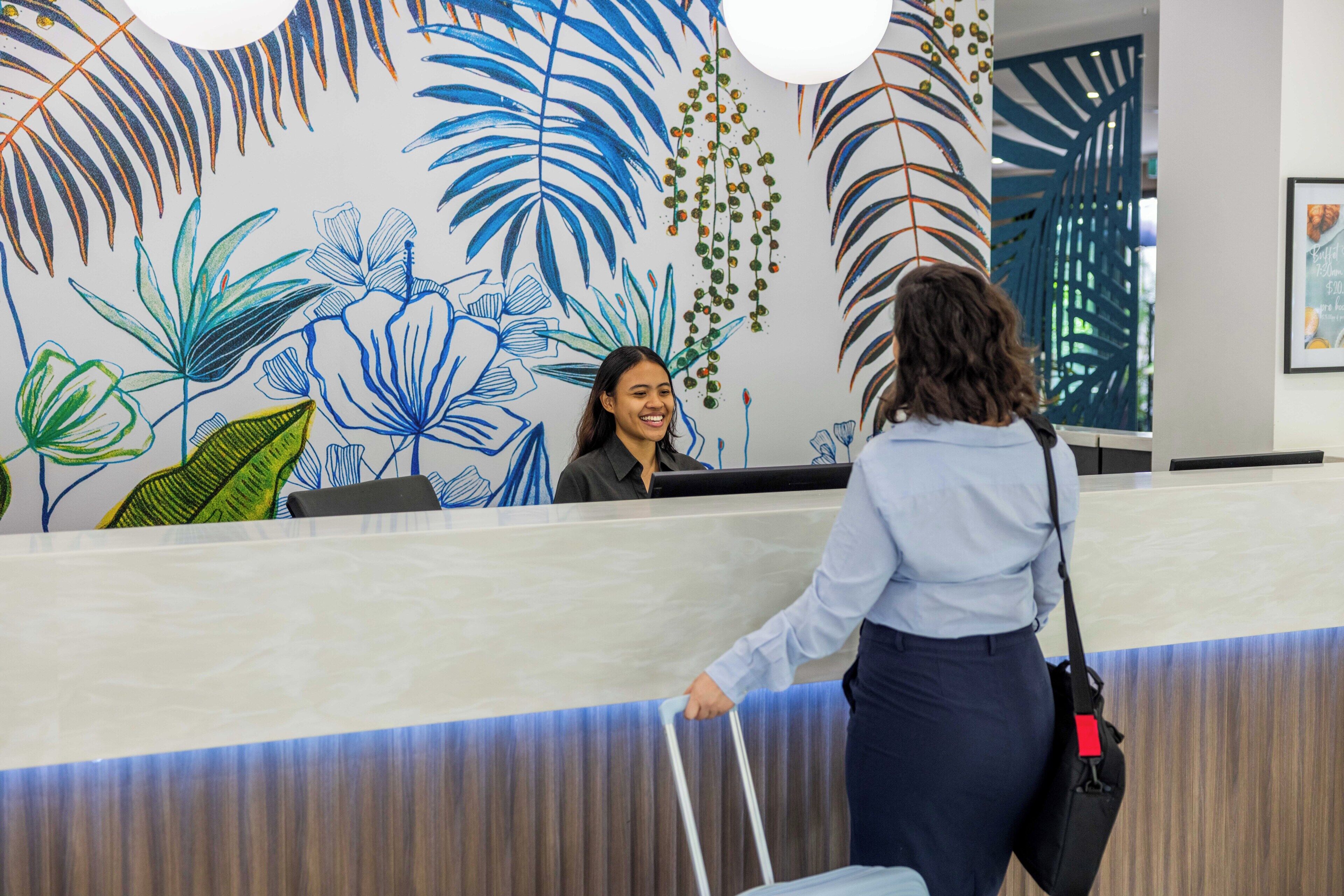 A reception desk with a mural of tropical leaves and a woman in a blue shirt and dark pants approaching.