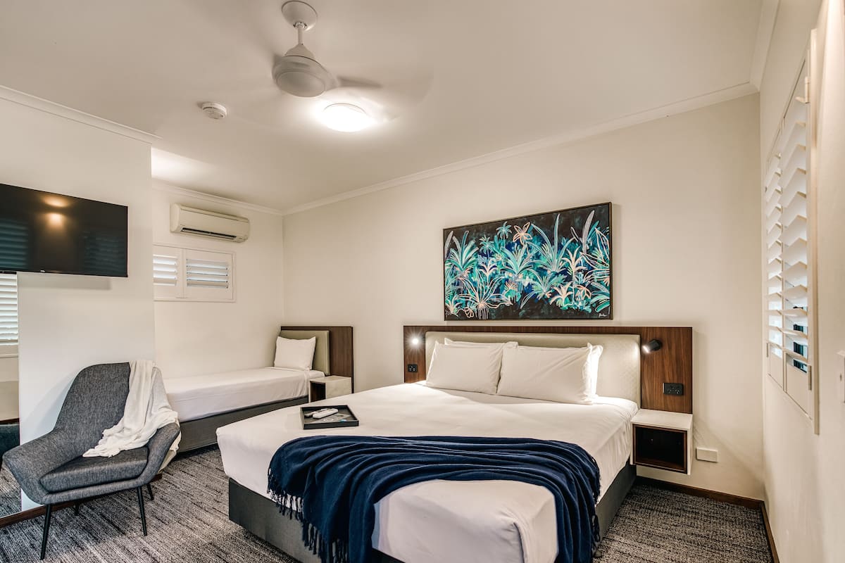 Una habitación de hotel con una cama grande, una cama más pequeña, una silla, un televisor de pantalla plana, un equipo de aire acondicionado y un ventilador de techo.