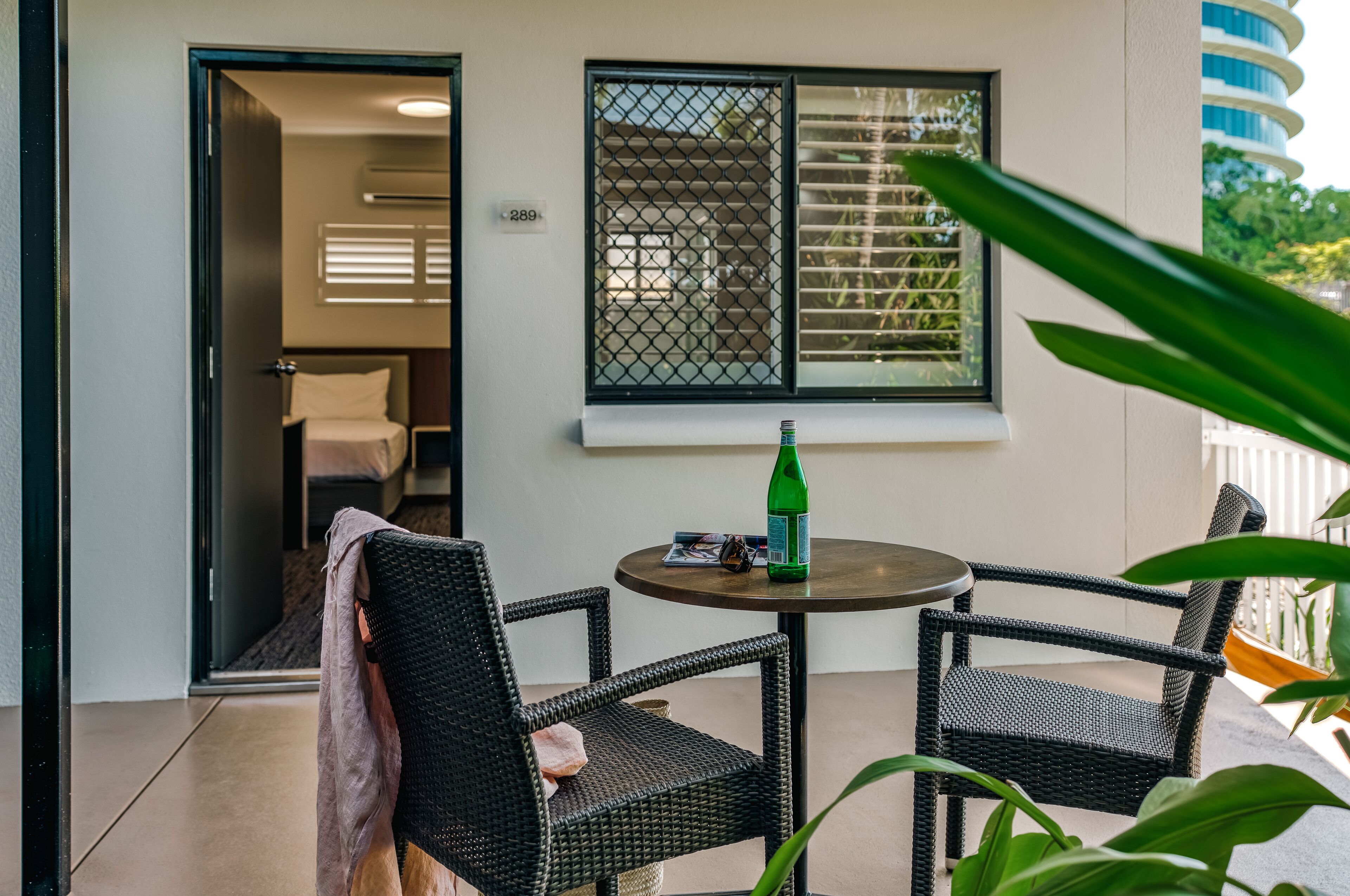 Ein Balkon mit Korbstühlen, einem Tisch mit einer Flasche und Sonnenbrille sowie einem Fenster mit Rollläden.