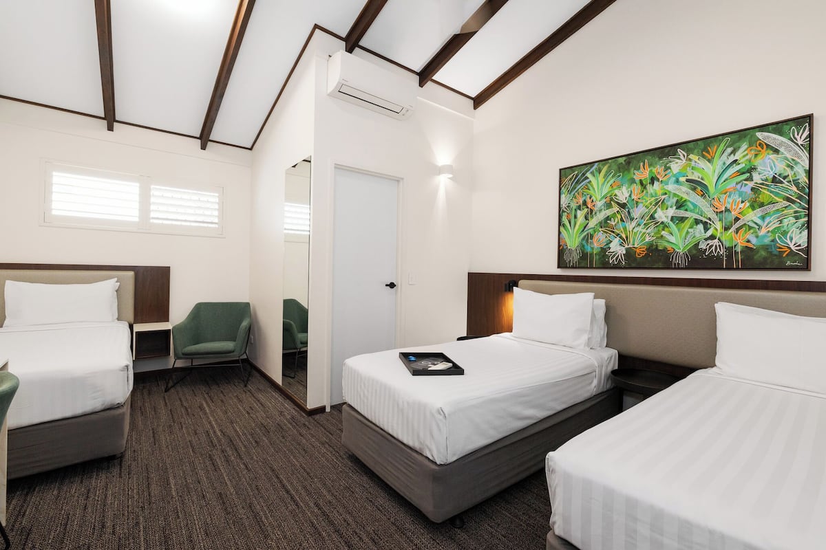 Habitación de hotel con dos camas, una silla verde, un cuadro en la pared y una puerta que da a otra habitación.