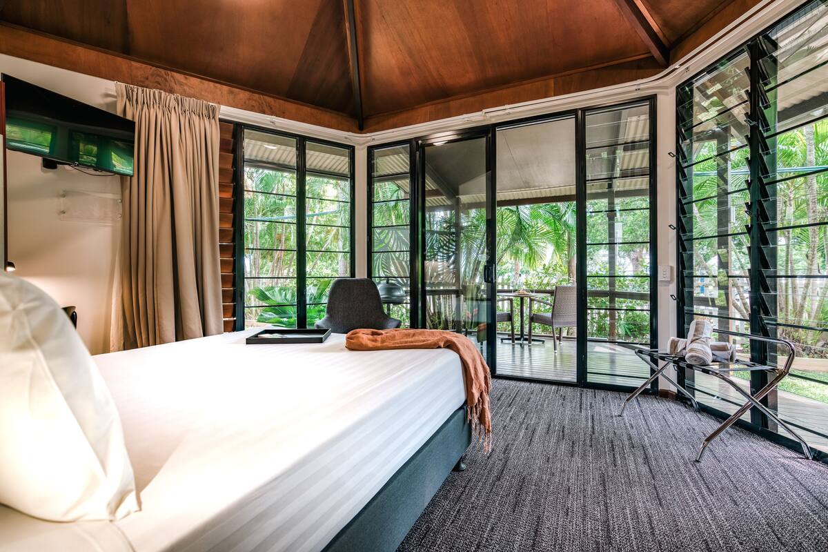 Un dormitorio con una cama grande, televisor y vistas a un balcón con sillas y vegetación.