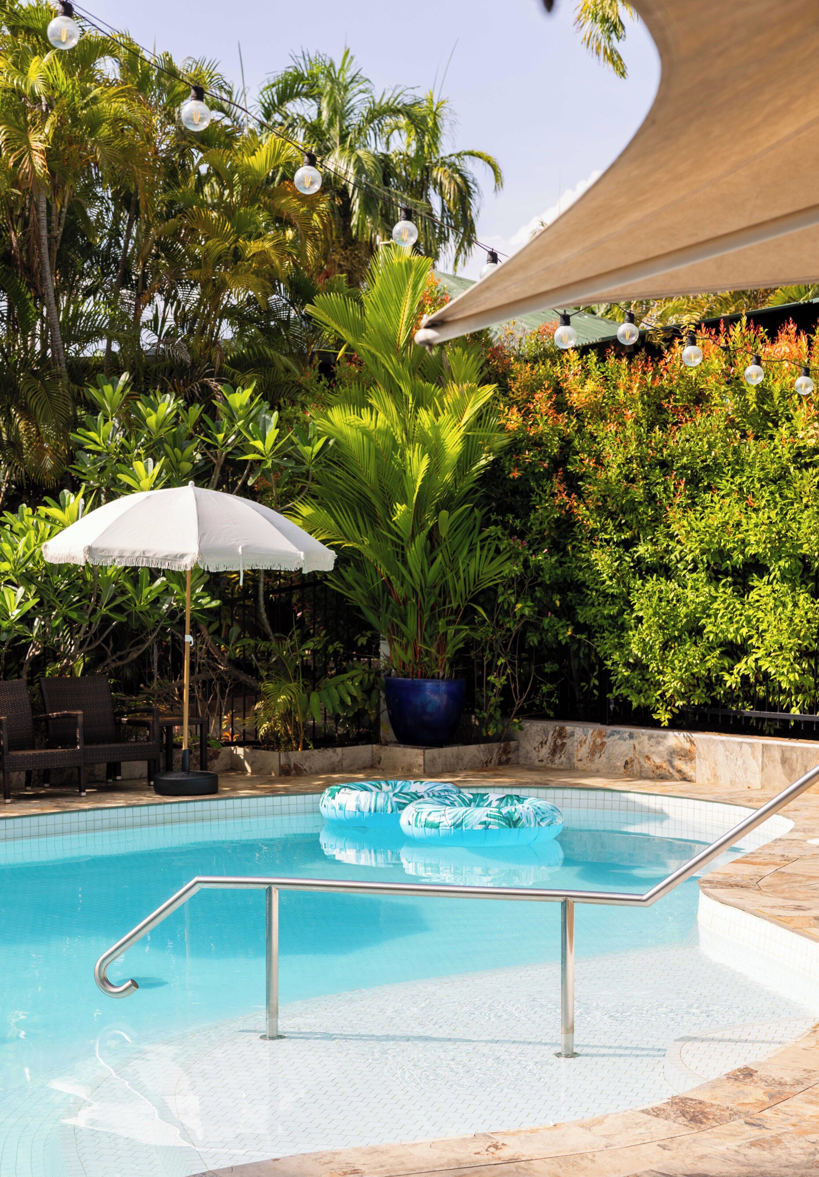 A swimming pool with a clear blue water, surrounded by lush greenery and a white umbrella.