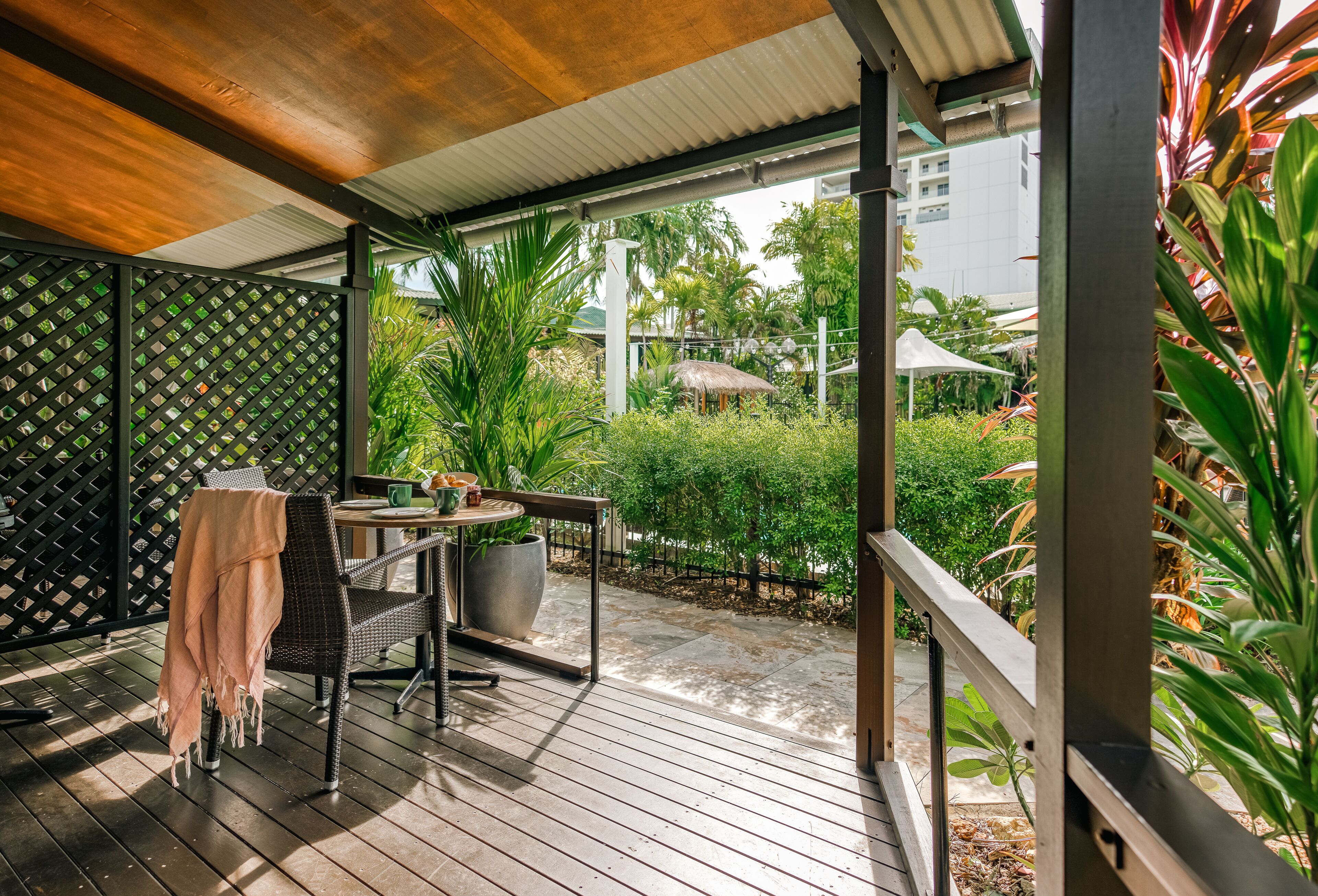 A covered outdoor seating area with a table and chairs, surrounded by greenery and a view of a residential area.