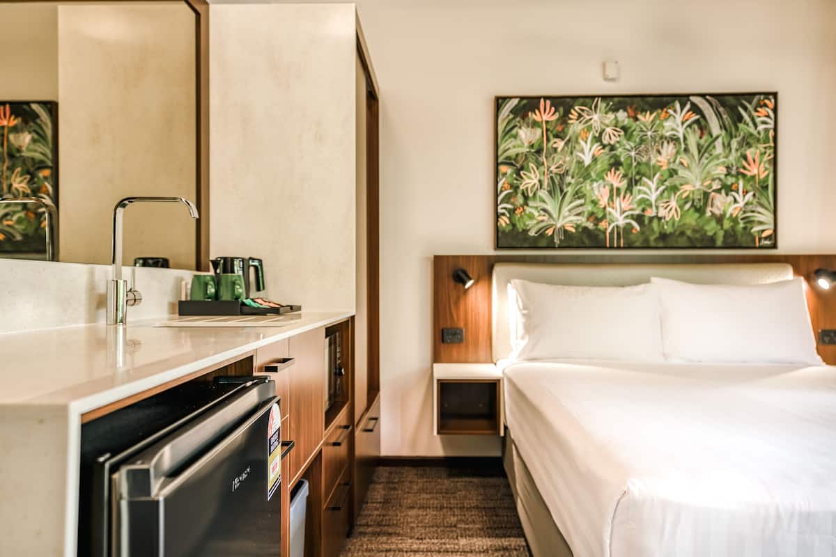Habitación de hotel con cama, escritorio con lavamanos, refrigerador, un cuadro en la pared y una mesita de noche con lámpara.