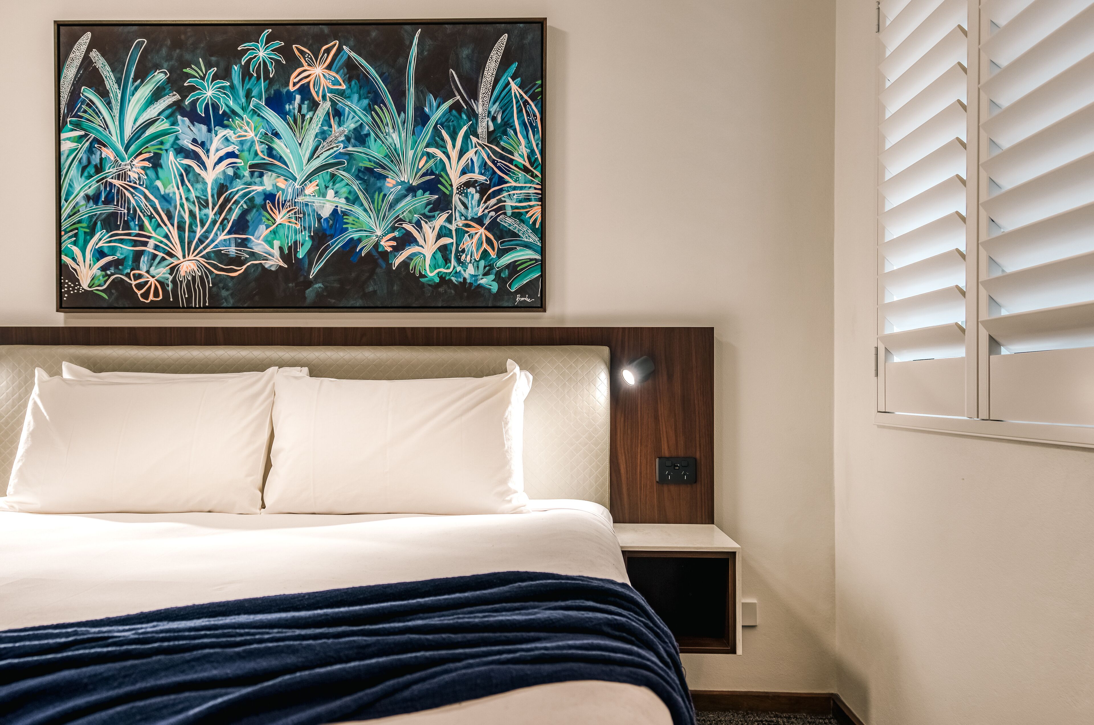 A hotel room with a bed, a nightstand, a wall-mounted artwork, and a window with blinds.