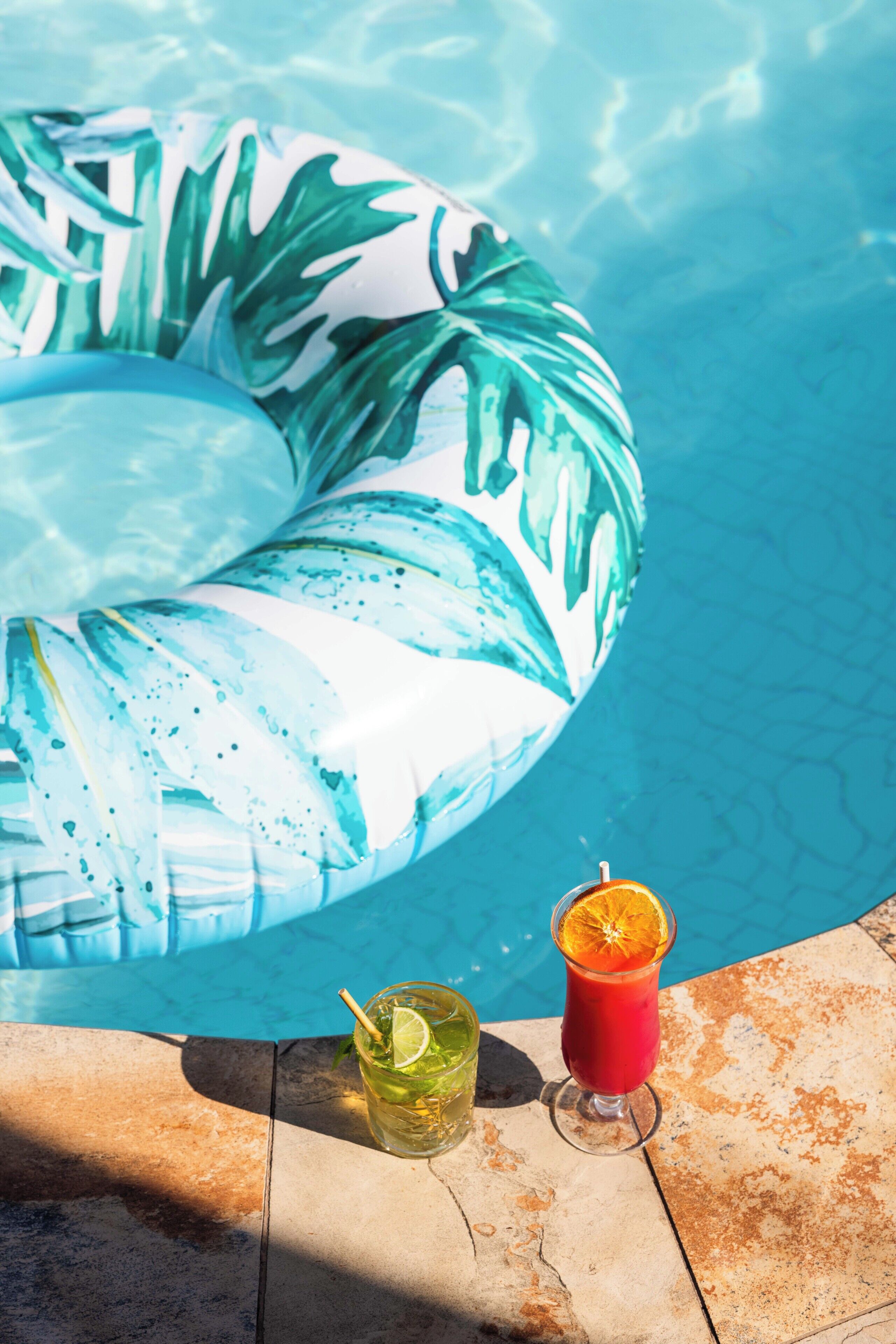 A pool with a large inflatable ring, a glass of red drink with an orange slice, and a glass of green drink with a straw and lime.