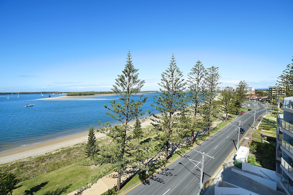 A coastal road with a beach and ocean view, surrounded by trees and buildings.