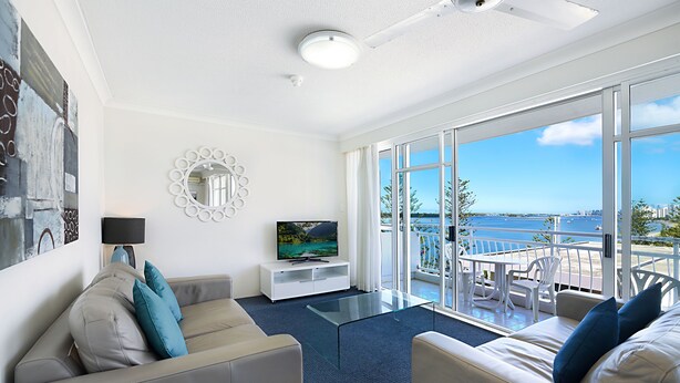 A modern living room with a sofa, a glass coffee table, and a television. There is a balcony with a view of the sea and buildings. - Atrium Resort (Biggera Waters)