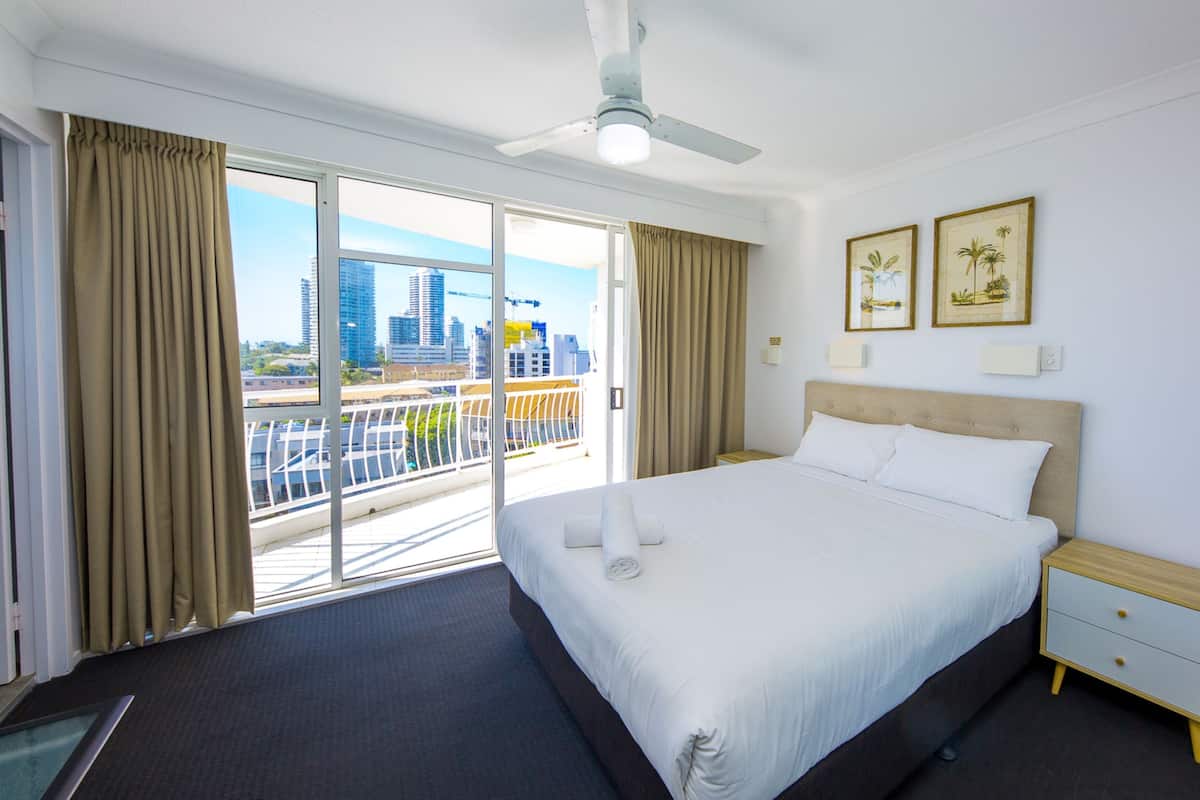 A hotel room with a large bed, a nightstand, a ceiling fan, and a view of the cityscape through the balcony doors.