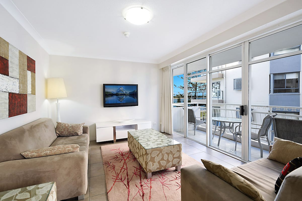 A modern living room with a sofa, a coffee table, and a TV. There is a balcony with a table and chairs.