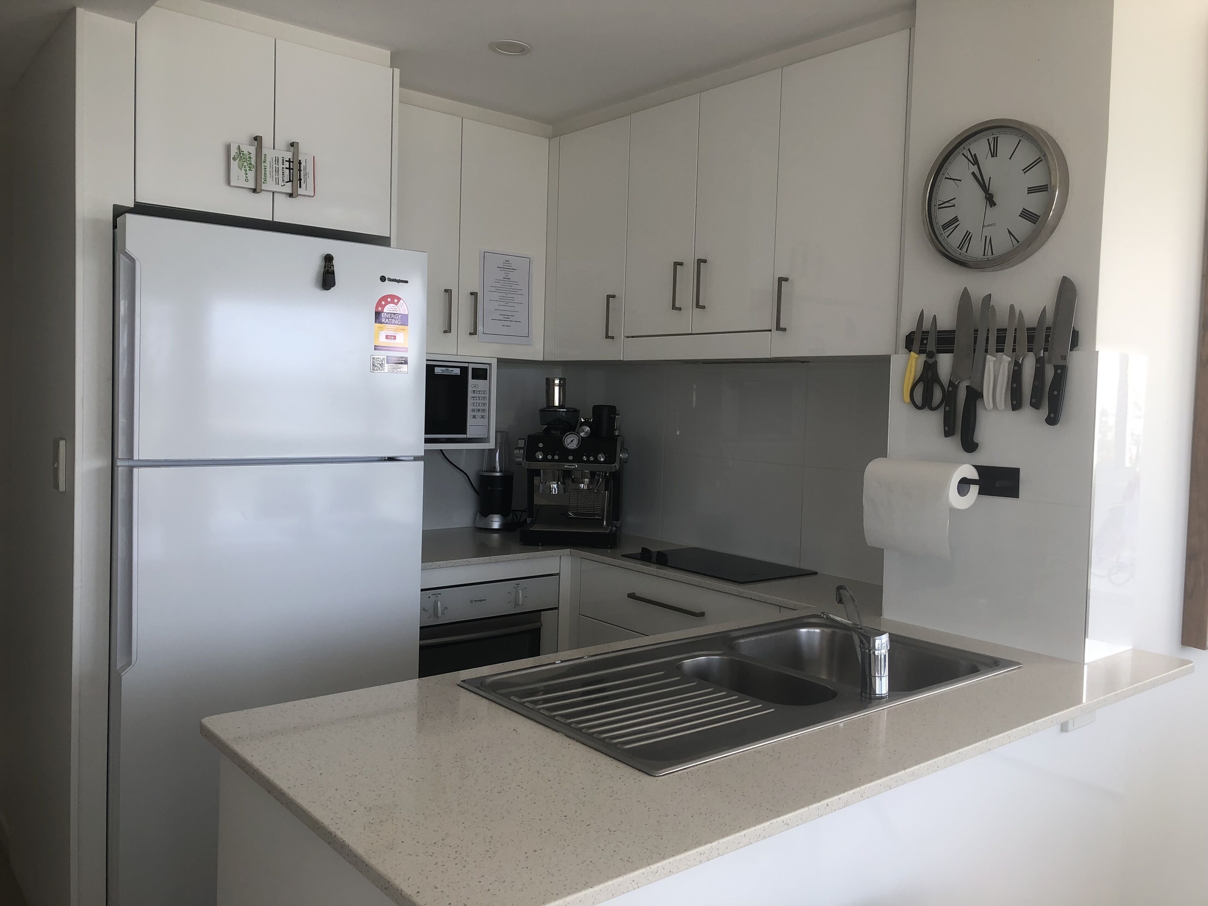 Una cocina moderna con armarios blancos, un refrigerador de acero inoxidable, un microondas, una máquina de espresso y un lavabo.