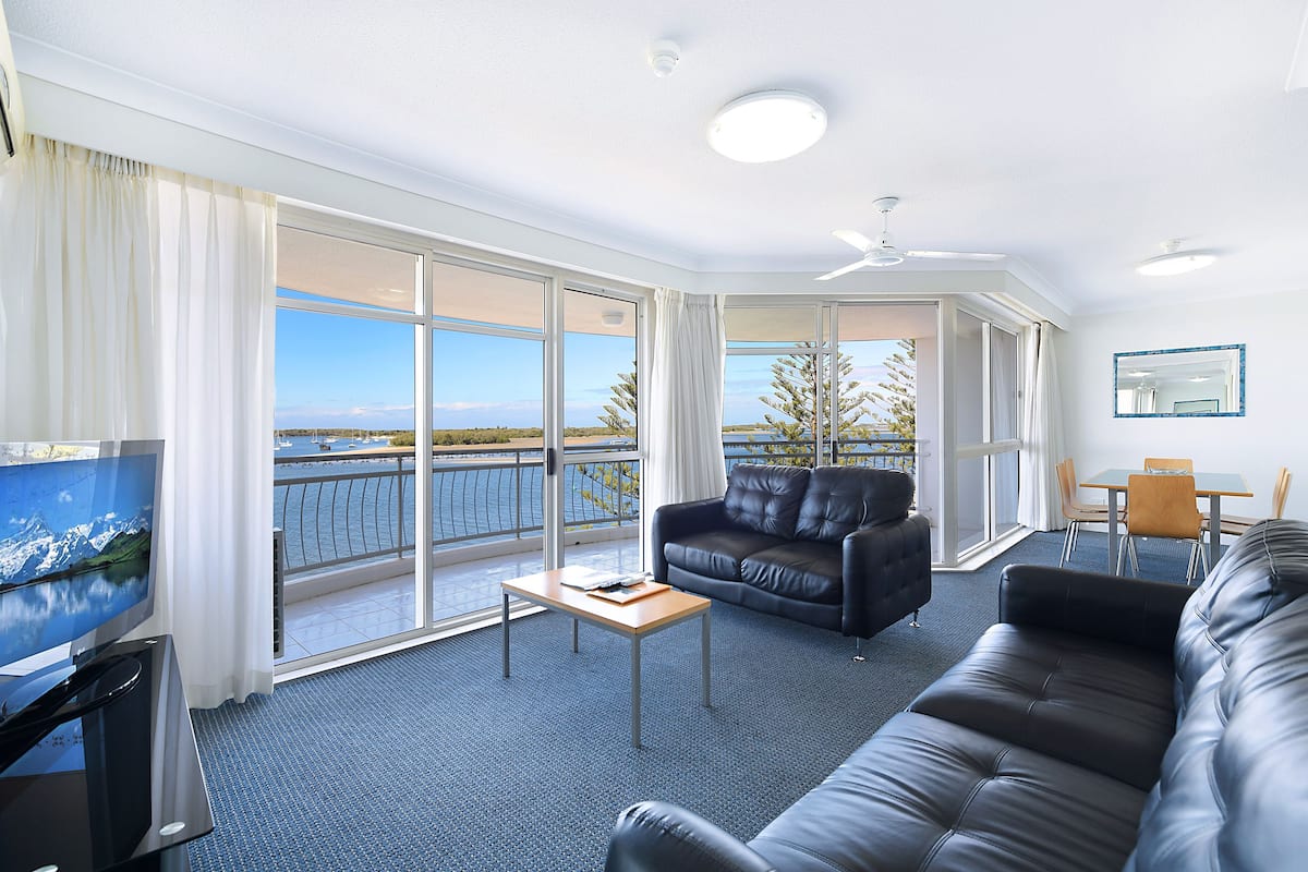 A modern hotel room with a large balcony, a leather sofa, a wooden coffee table, and a flat-screen TV.
