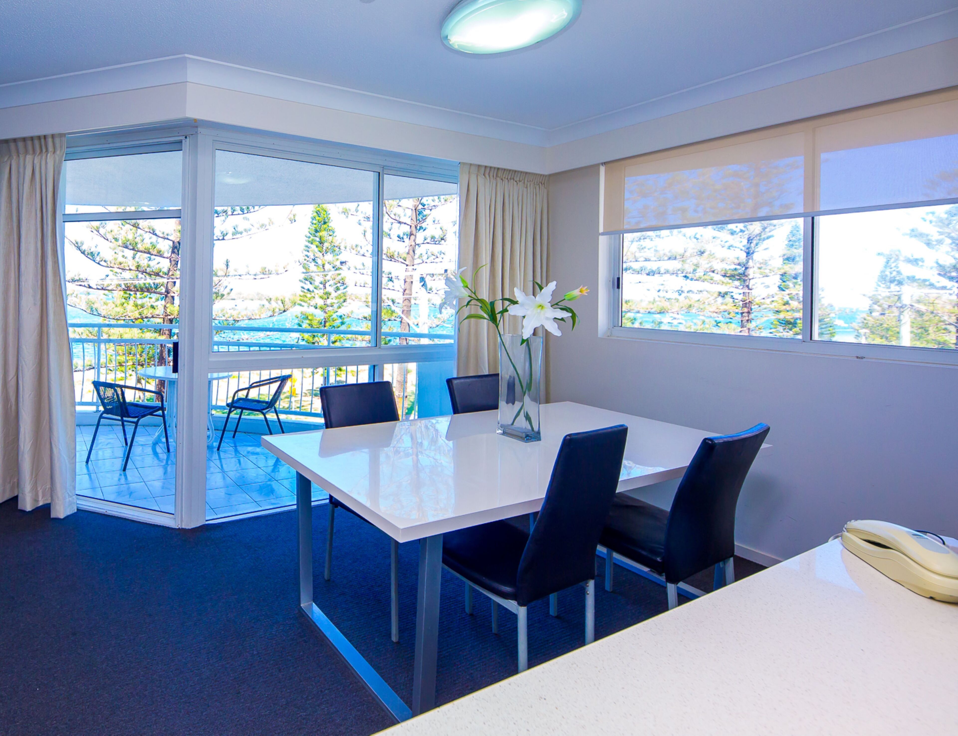 Ein Zimmer mit einem weißen Tisch, vier schwarzen Stühlen, einer Vase mit weißen Blumen und einem Fenster mit Blick auf Bäume und einen blauen Himmel.