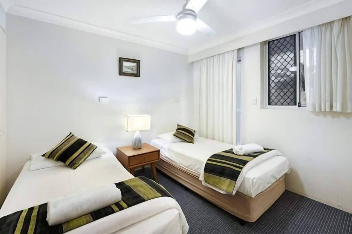 A hotel room with two single beds, a nightstand with a lamp, a ceiling fan, a window with curtains, and a picture on the wall.