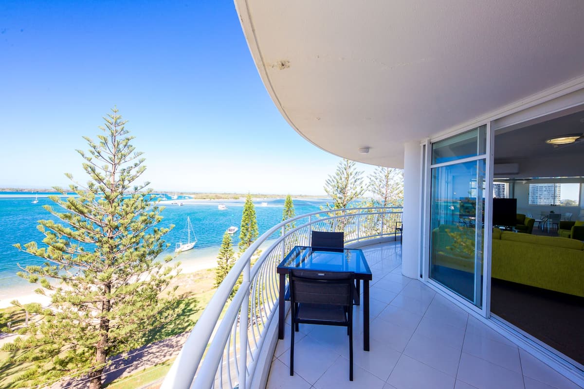 A balcony with a glass door, a table with a chair, and a view of the sea and trees.