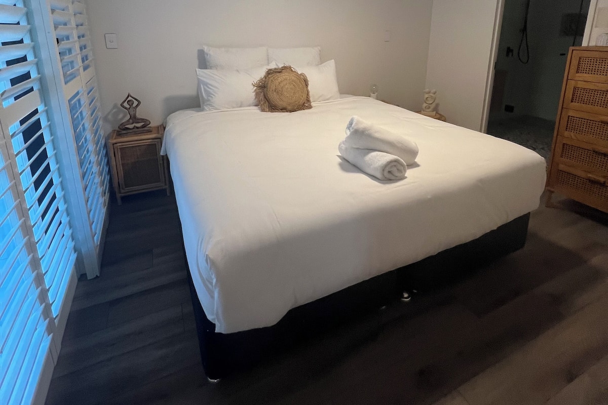 A neatly made bed with white linens, a round throw pillow, and a folded towel on the foot of the bed. There is a small bedside table with a lamp and a wall clock. The room has a wooden floor and a door leading to a bathroom with a shower.
