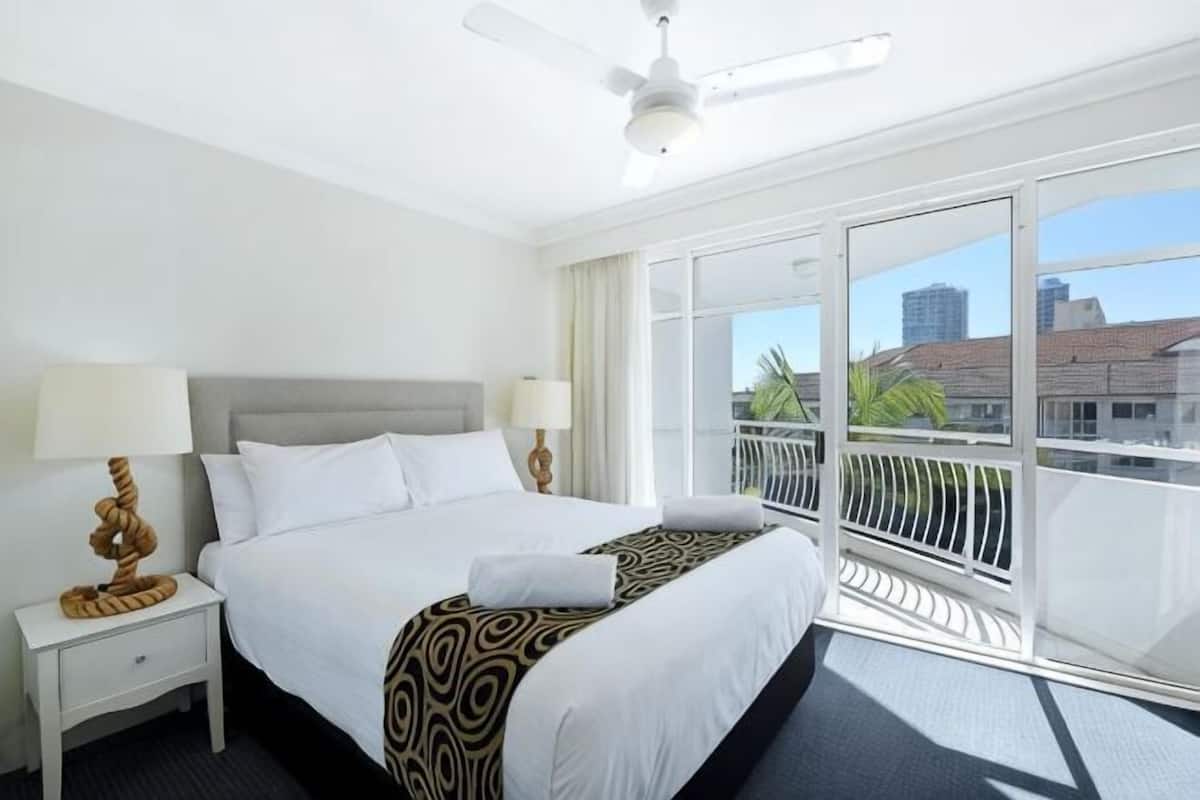 A hotel room with a large bed, two bedside tables, a ceiling fan, and a balcony with a view of buildings and palm trees.