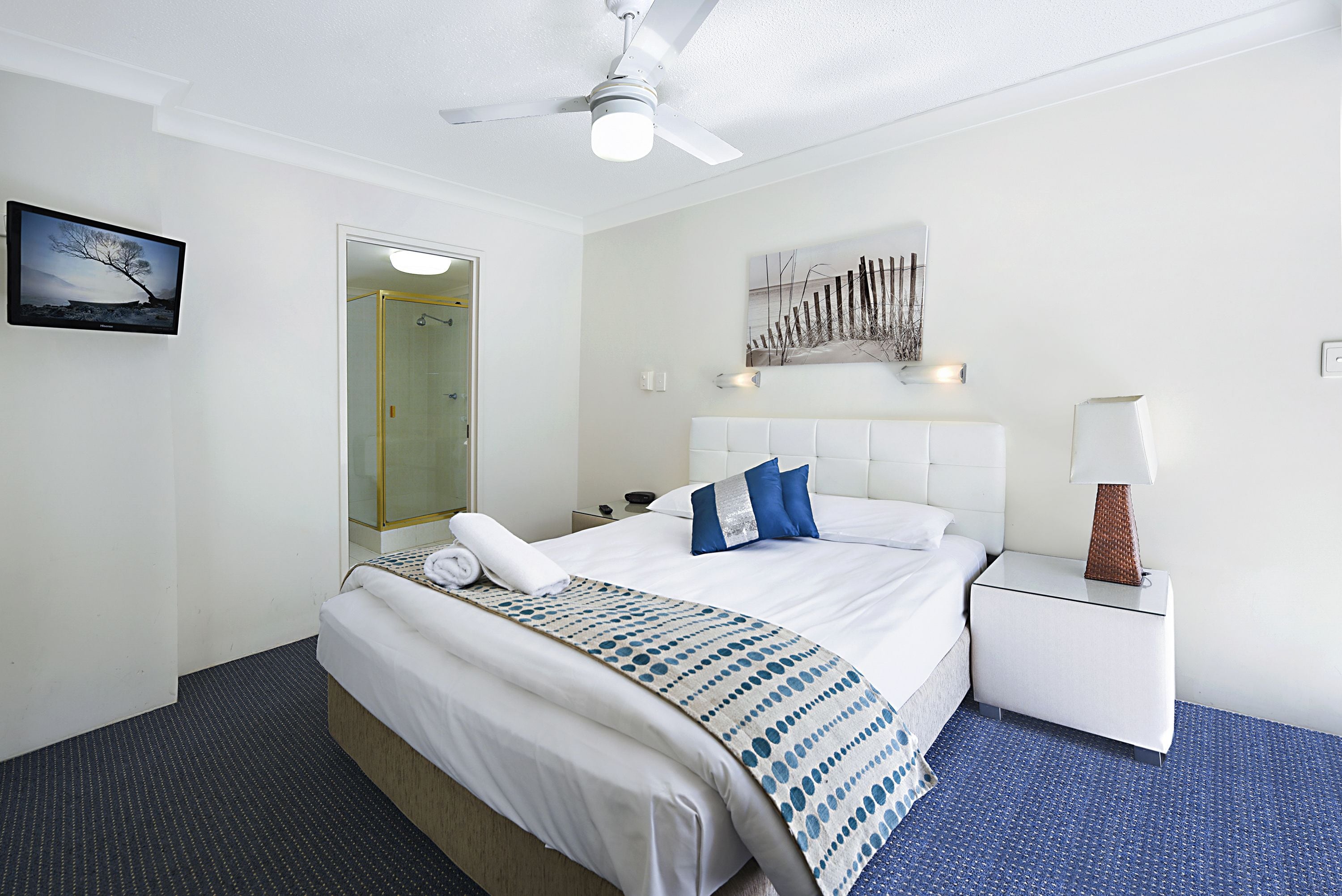 Habitación de hotel con cama, mesita de noche, lámpara, ventilador de techo y un cuadro en la pared.