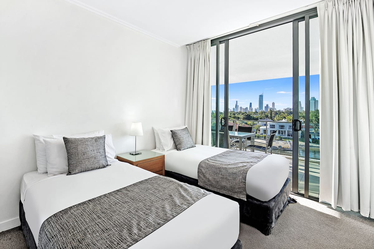 Ein Hotelzimmer mit zwei Betten, einem Nachttisch, einer Lampe und einem großen Fenster mit Blick auf die Stadt.