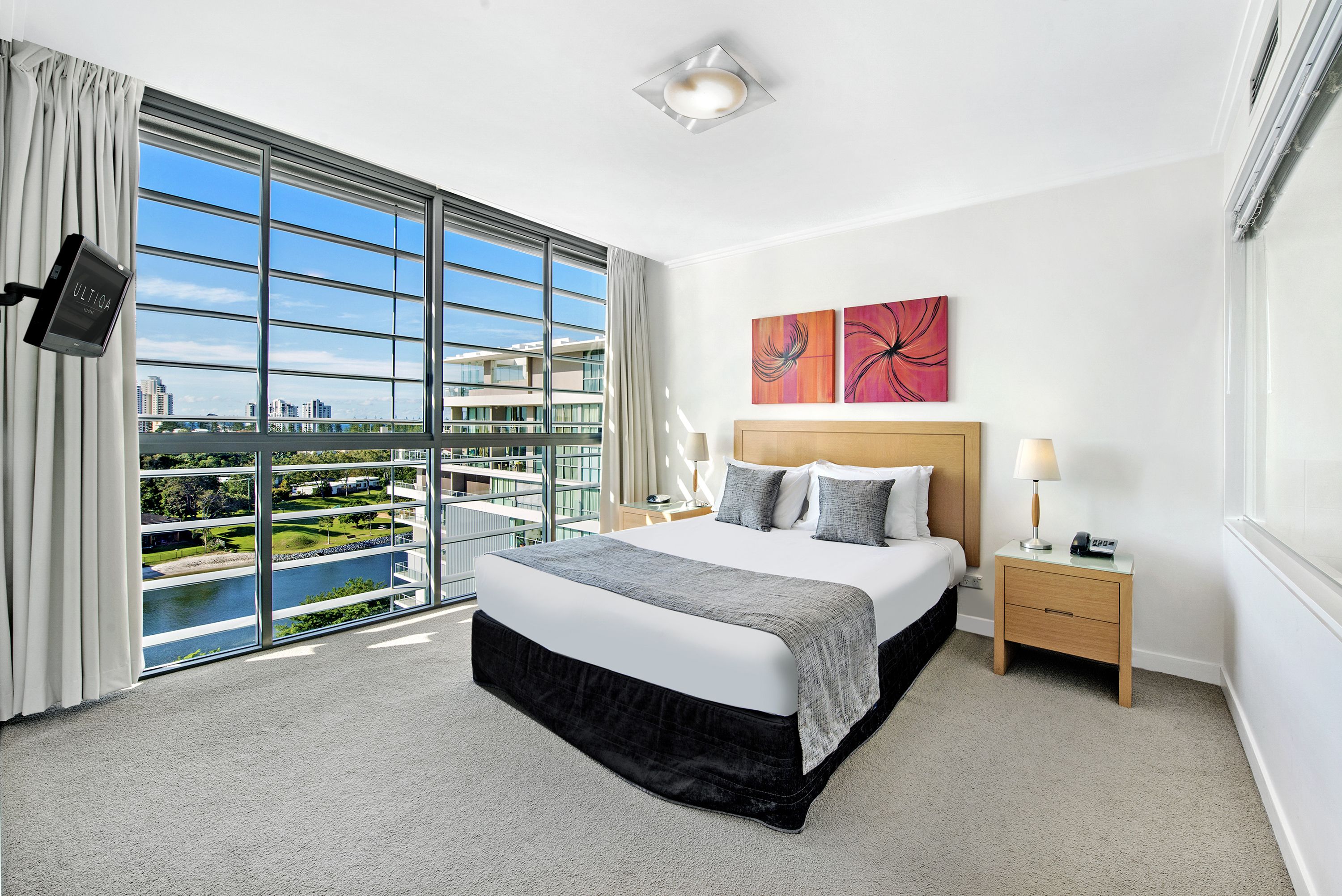 A modern hotel room with a large bed, a nightstand, a TV, and a view of a cityscape and water through floor-to-ceiling windows.