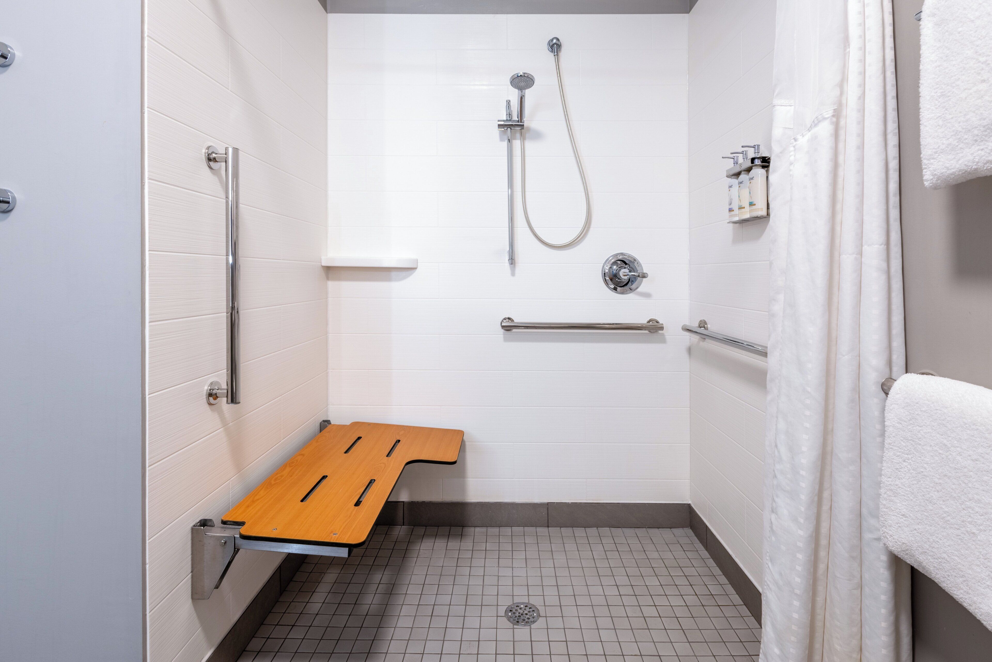 Une salle de bain équipée d’une douche, de barres d’appui, d’un banc en bois et d’un porte-serviettes.