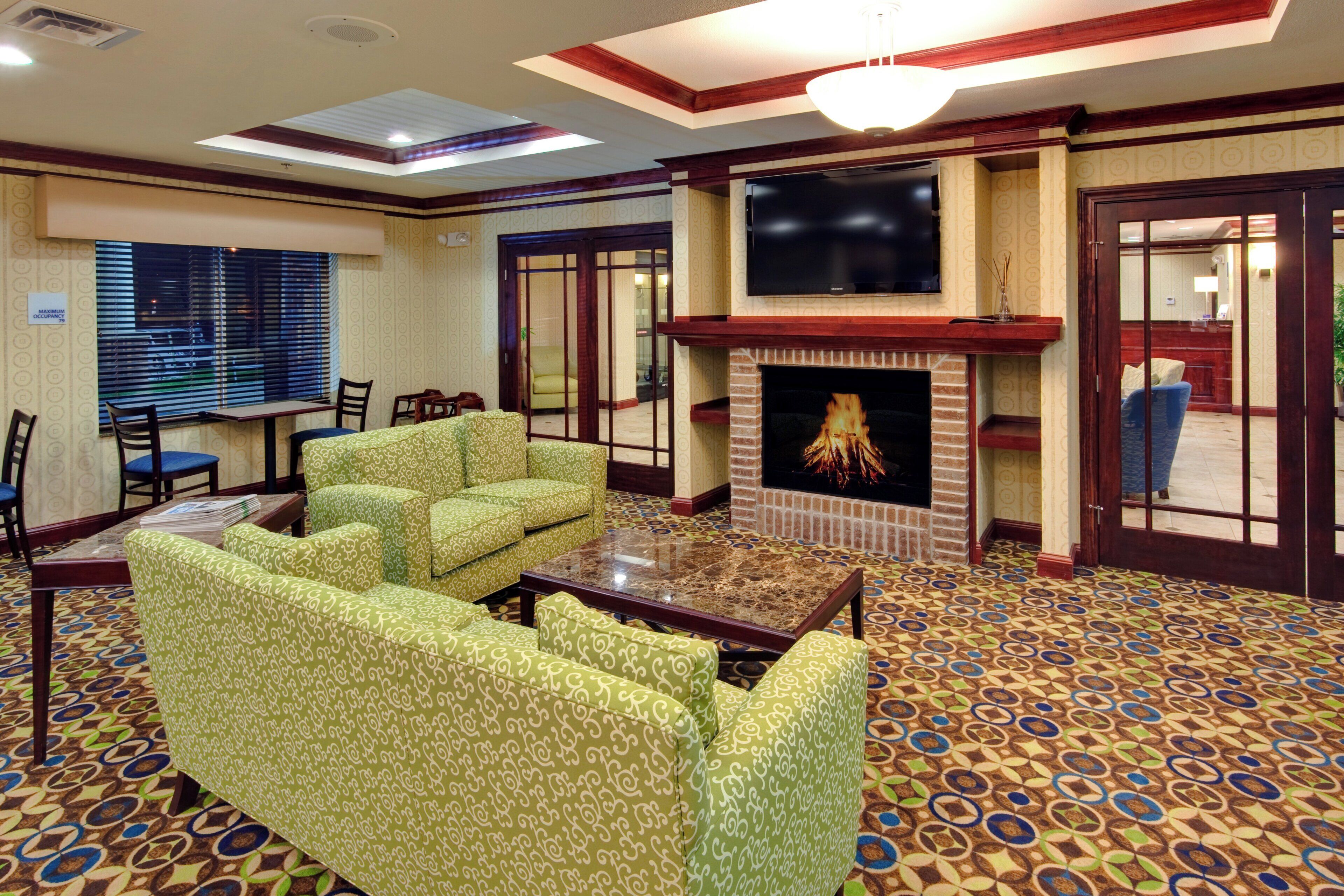 A hotel lobby with patterned carpet, green patterned sofas, a fireplace, a flat-screen TV, and a reception desk.