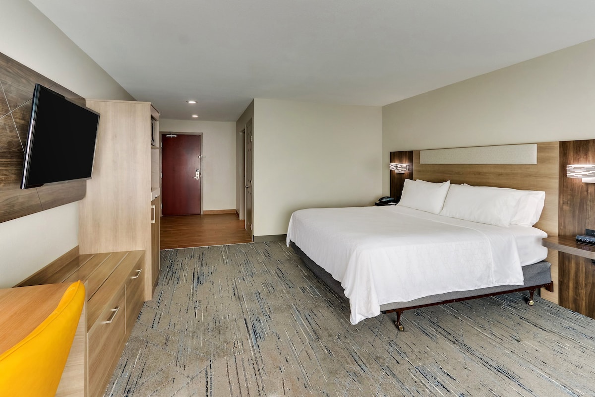 A hotel room with a large bed, a flat-screen TV mounted on the wall, a wooden desk, and a bright yellow chair.