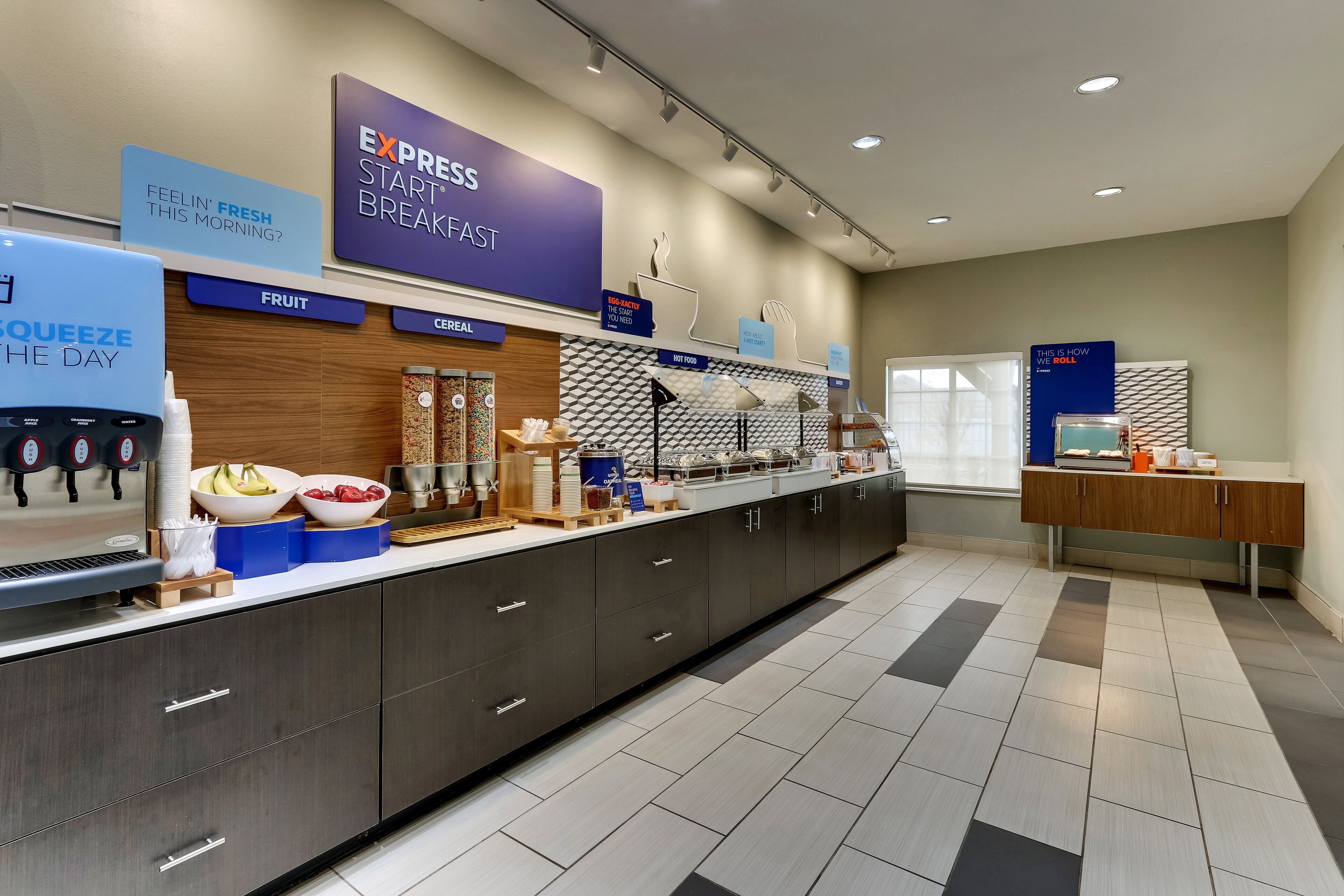 A modern hotel breakfast area with a self-serve station, fruit and cereal options, and a sign promoting a fresh start to the day.