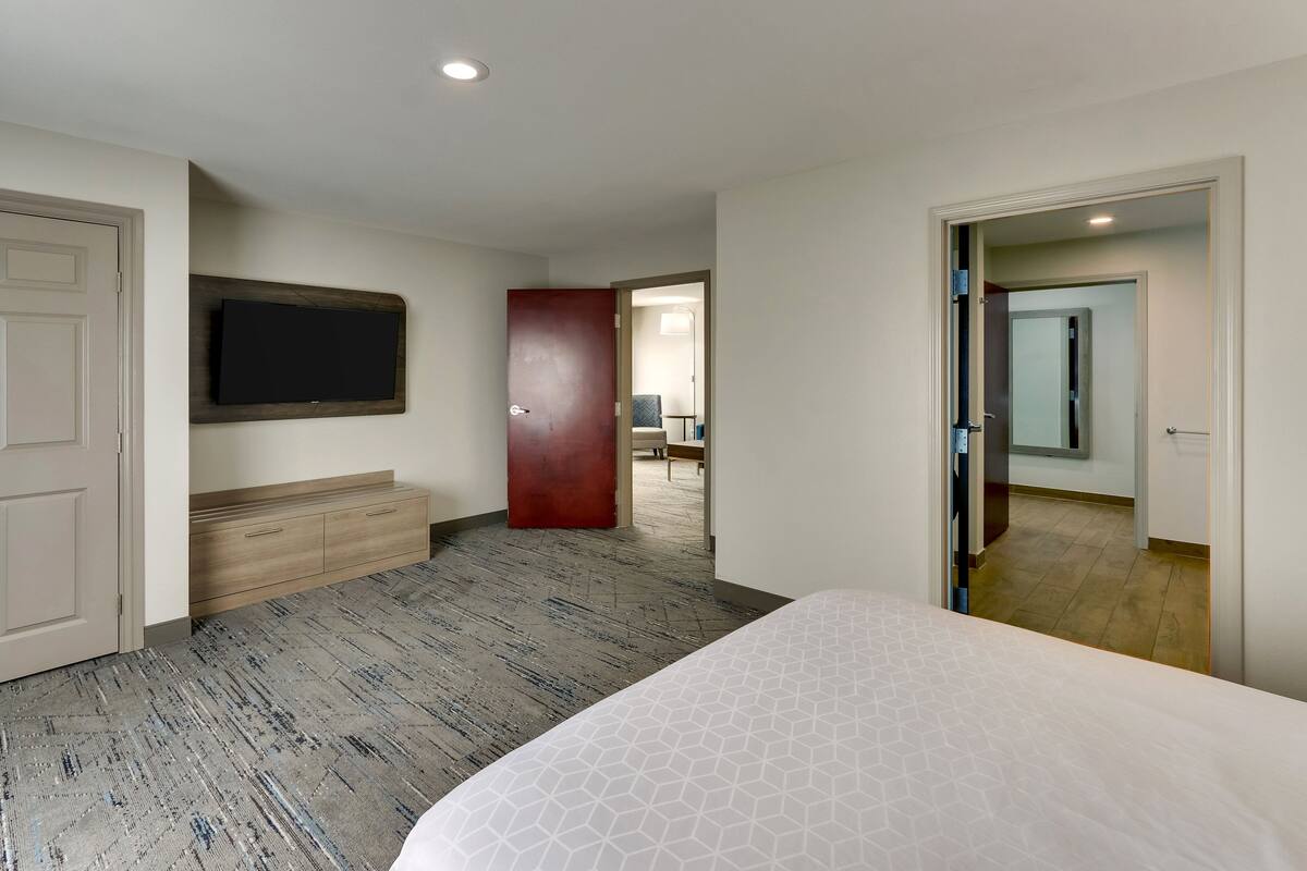 A hotel room with a large bed, a flat-screen TV mounted on the wall, a wooden dresser, and a red door leading to another room.