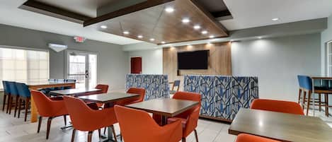 A modern dining area with wooden tables and orange chairs, a patterned wall, and a flat-screen TV mounted above a counter.