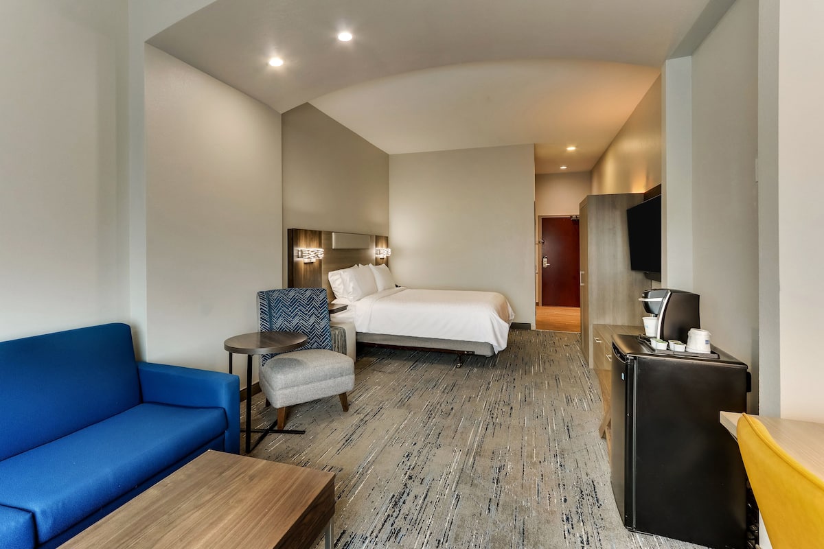 A hotel room with a bed, a blue sofa, a small table, a chair, a desk with a flat-screen TV, and a refrigerator.