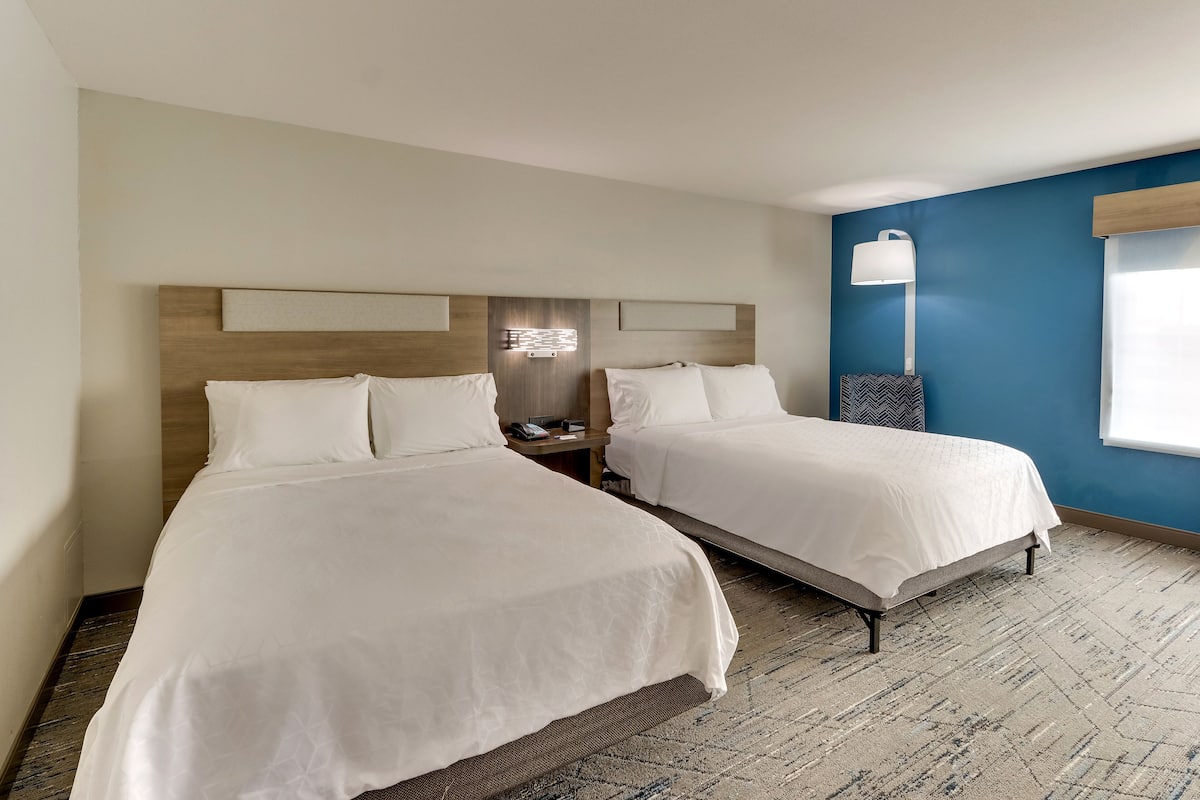 A hotel room with two beds, a nightstand, a lamp, and a window with blinds.