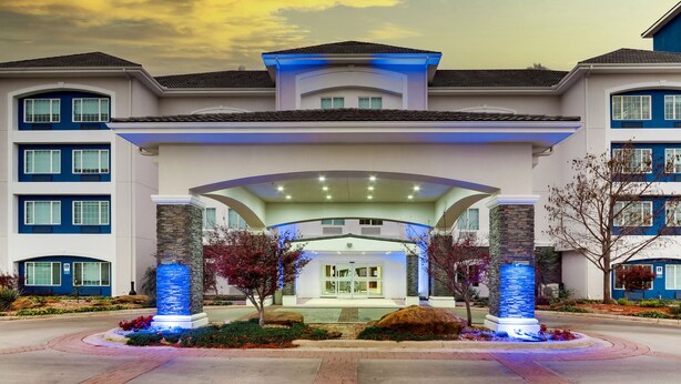 A hotel with a modern architectural design, featuring a central entrance with a glass facade, stone pillars, and a landscaped garden with a water feature.