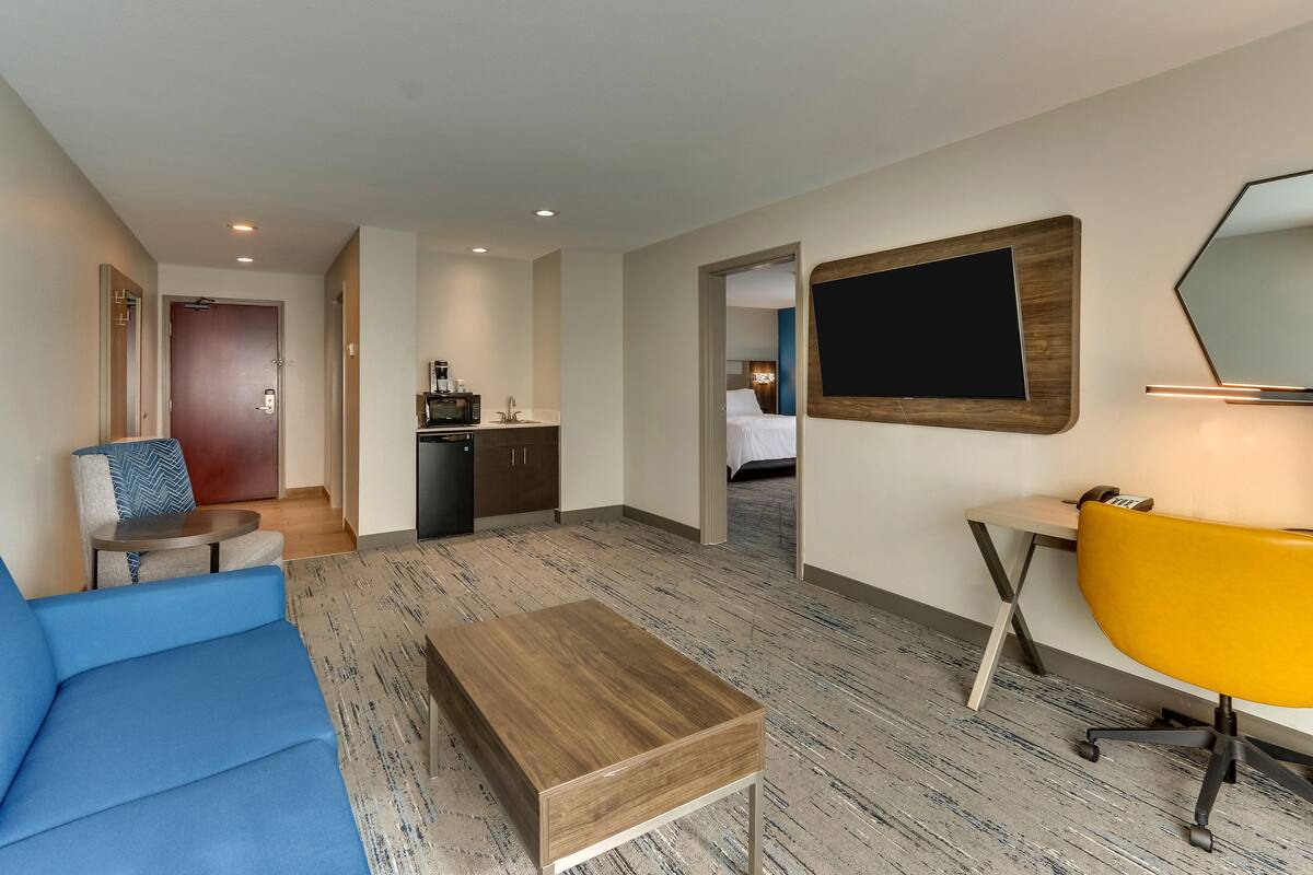 A modern hotel room with a blue sofa, a wooden coffee table, a yellow chair, a desk with a lamp, and a large mirror on the wall.