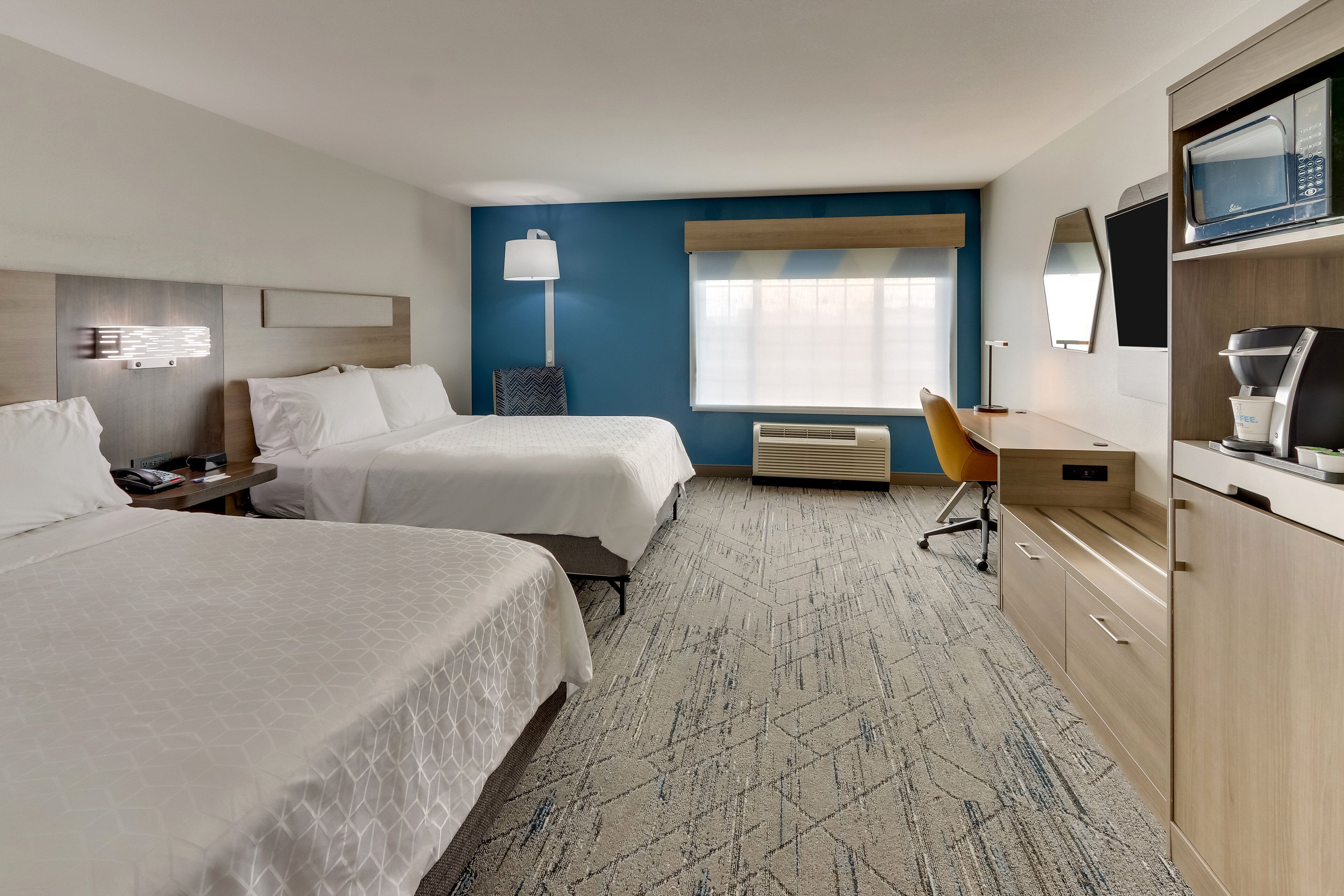 Habitación de hotel con dos camas, un escritorio, una silla, microondas, cafetera y un ventanal grande con persianas.