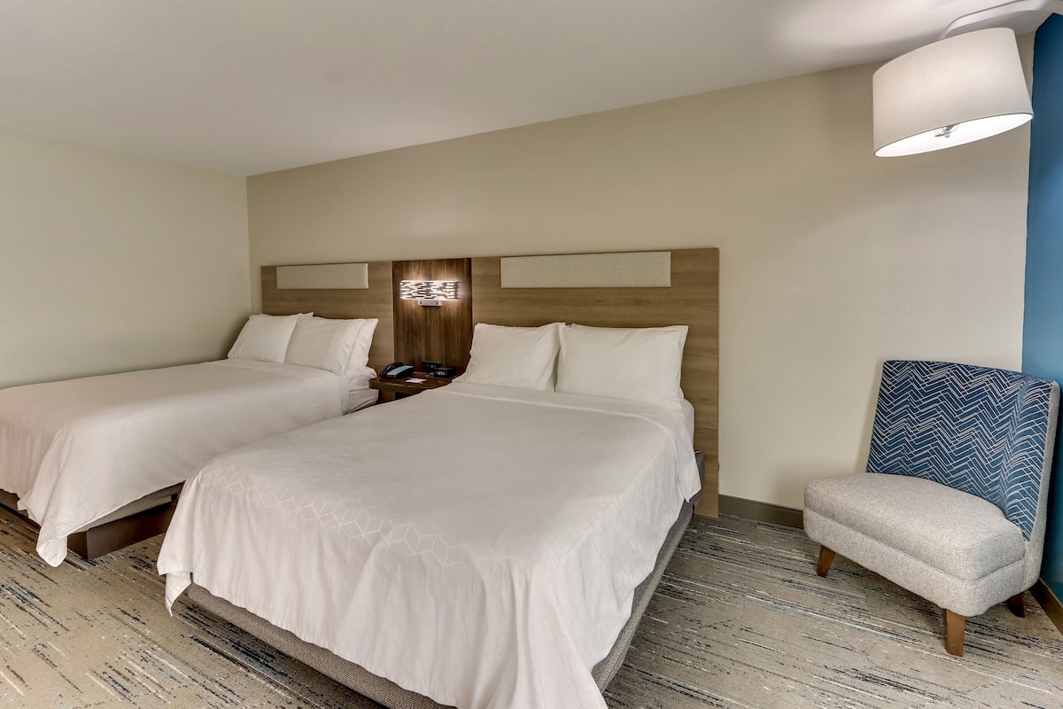 A hotel room with two beds, a chair, a lamp, and a wall-mounted phone.