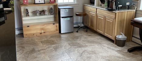 Una cocina con refrigerador, cafetera y ventana. Hay premios enmarcados en la pared y un reloj sobre la ventana.